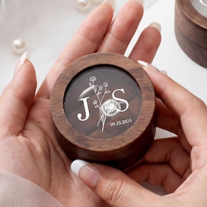 May include: Two round dark wood ring boxes with clear lids. One box is held, showing white floral line art, the letters J and S, and the date 09.23.2025. The second box sits nearby, featuring white floral line art and the letters J and A.