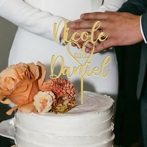 Pode incluir: Um topo de bolo dourado com os nomes "Nicole" e "Daniel" em escrita elegante, conectados por um coração com a data "15.27.23". O topo está colocado em um bolo branco decorado com flores pêssego e rosa. As mãos de um casal são visíveis ao fundo.