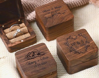 May include: Four wooden ring boxes, three closed and one open, displaying a silver ring. The closed boxes are engraved with names and dates, and decorative elements. The open box has a burlap interior.
