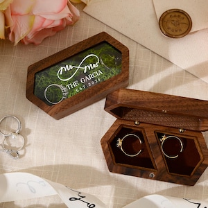 Puede incluir: Dos joyeros de madera para anillos, uno abierto y otro cerrado, con anillos de boda en su interior. La caja cerrada tiene el texto "mr & mrs THE GARCIA EST. 2025". La caja abierta muestra dos anillos en compartimentos separados.