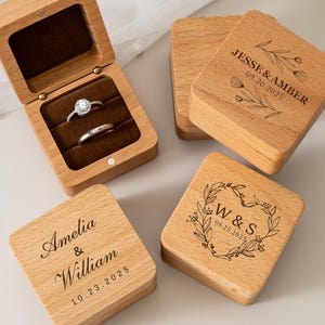 May include: Four square wooden ring boxes with hinged lids. One box is open, revealing a dark brown velvet interior holding two silver rings. The closed boxes feature laser-engraved names, dates, and floral designs including Jesse & Amber, Amelia & William, and W & S.