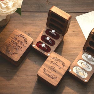May include: Two rectangular dark wood ring boxes with hinged lids. One box is closed, showing engraved text "Mr. & Mrs. Williams Sep 15.2025" with leaf accents. The other box is open, displaying three silver rings with white stones in red velvet slots.