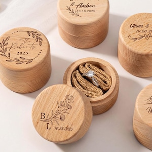 Könnte beinhalten: Eine Auswahl personalisierter Holz-Ringboxen mit Gravuren von Namen, Daten und floralen oder Bergmotiven. Eine Box ist geöffnet und zeigt einen Ring. Die Boxen sind aus hellem Holz.