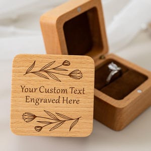 May include: A wooden ring box with a hinged lid, open to reveal a ring on a brown velvet interior. The box is engraved with floral designs and the text "Your Custom Text Engraved Here".