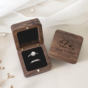 May include: A dark brown wooden ring box with a black velvet interior containing two silver rings. The lid features an engraved mountain landscape, the names Henry + Olivia, and the date 08.25.2025. The box is square with a hinged lid.