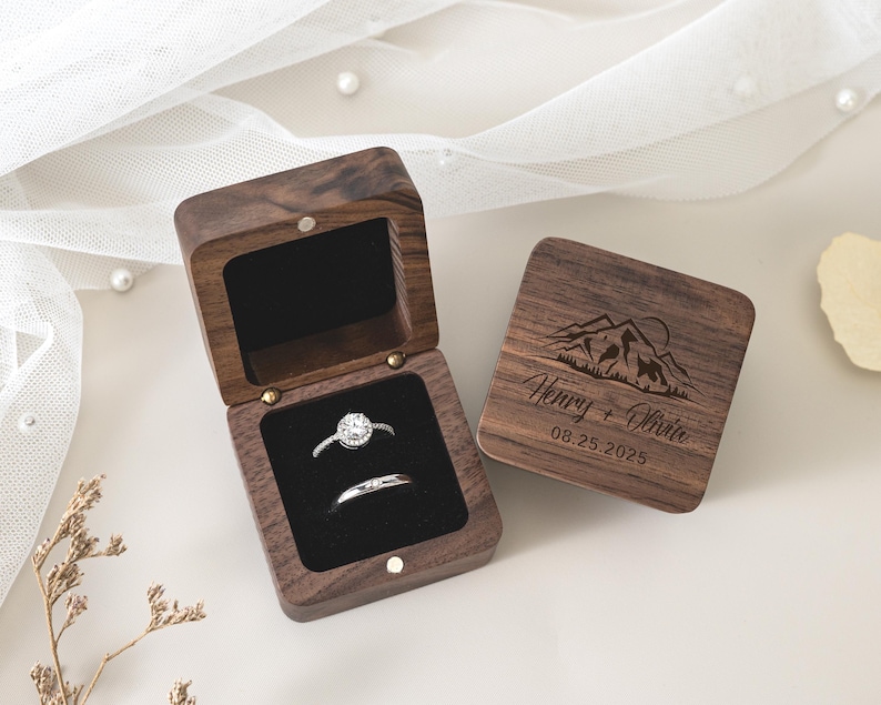 May include: A dark brown wooden ring box with a black velvet interior containing two silver rings. The lid features an engraved mountain landscape, the names Henry + Olivia, and the date 08.25.2025. The box is square with a hinged lid.