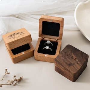 May include: Two square wooden ring boxes with black velvet inserts. One light wood box is open, holding two silver rings, and engraved with "Henry + Olivia 08.25.2026" and a mountain graphic. A closed dark wood box is engraved with "W & S 08.25.2025".