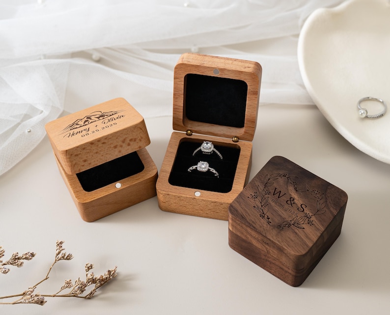 May include: Two square wooden ring boxes with black velvet inserts. One light wood box is open, holding two silver rings, and engraved with "Henry + Olivia 08.25.2026" and a mountain graphic. A closed dark wood box is engraved with "W & S 08.25.2025".