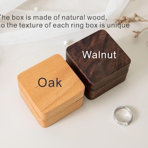 May include: Two square ring boxes made of natural wood, one light-coloured labelled Oak and one dark-coloured labelled Walnut. The text states: The box is made of natural wood, so the texture of each ring box is unique. A silver ring sits nearby.