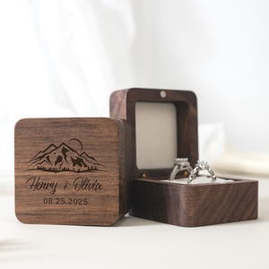 May include: A dark brown wooden ring box with a hinged lid, featuring an engraved mountain range graphic, the names Henry + Olivia, and the date 08.25.2025. The open box interior is lined with white fabric and holds two silver-toned rings.