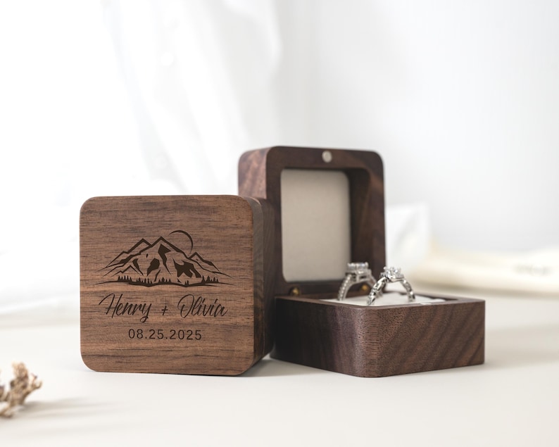 May include: A dark brown wooden ring box with a hinged lid, featuring an engraved mountain range graphic, the names Henry + Olivia, and the date 08.25.2025. The open box interior is lined with white fabric and holds two silver-toned rings.