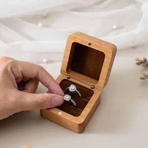 May include: A rectangular wooden ring box with a hinged lid and a dark brown velvet interior lining. The box contains two silver-toned rings, each featuring a central square-cut stone surrounded by a halo of smaller clear stones.