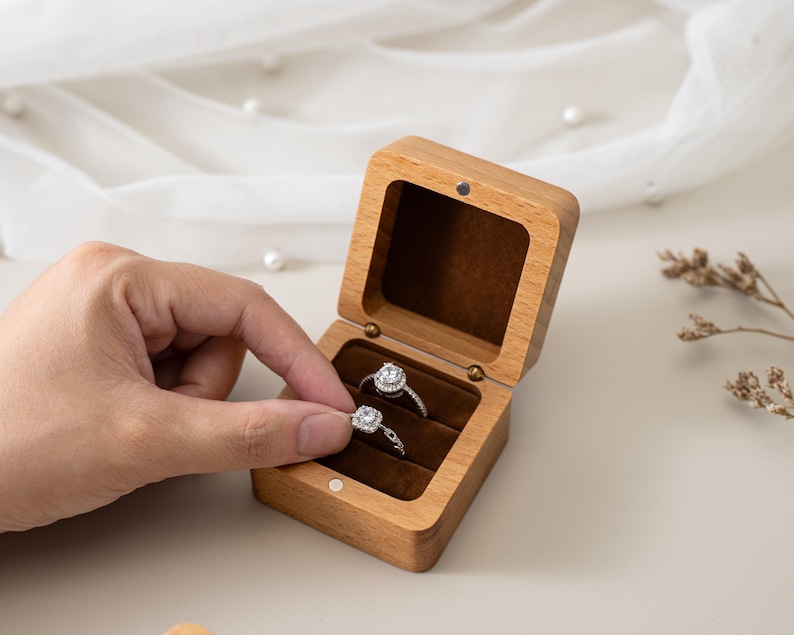 May include: A rectangular wooden ring box with a hinged lid and a dark brown velvet interior lining. The box contains two silver-toned rings, each featuring a central square-cut stone surrounded by a halo of smaller clear stones.
