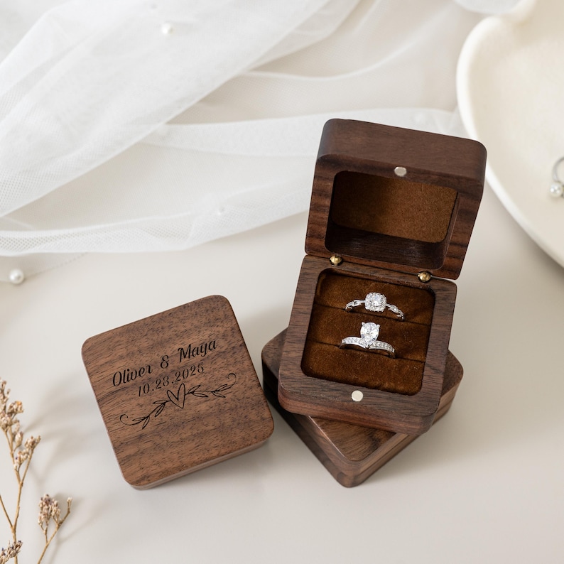 May include: A square dark brown wooden ring box with a hinged lid. The lid is engraved with the text Oliver & Maya, the date 23.10.2025, and a heart design. The open box features a brown velvet interior holding two silver rings with stones.