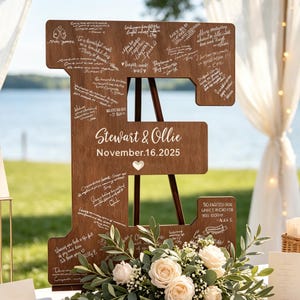 May include: A large wooden letter E guestbook sign featuring a dark brown finish, white printed text reading Stewart & Ollie November 16, 2025, and a small heart icon. The surface is covered in numerous handwritten messages in white ink.