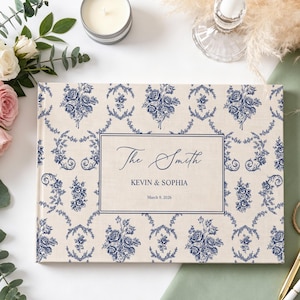 Op de afbeelding: Een rechthoekig fotoalbum met een crèmekleurige stoffen kaft voorzien van een blauw bloemmotief in toile-stijl. Een centraal rechthoekig kader bevat de tekst The Smith, KEVIN & SOPHIA en de datum March 9, 2026, gedrukt in donkerblauwe inkt.