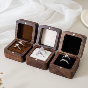 May include: Three square dark wood ring boxes with hinged lids. The left box has a brown velvet interior, the centre box has a white velvet interior, and the right box has a black velvet interior. Each box holds silver rings with clear stones.