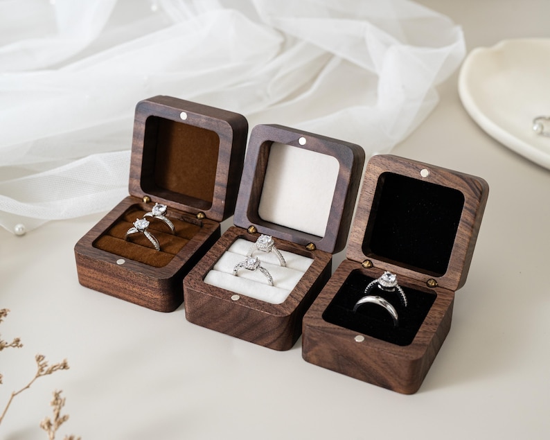 May include: Three square dark wood ring boxes with hinged lids. The left box has a brown velvet interior, the centre box has a white velvet interior, and the right box has a black velvet interior. Each box holds silver rings with clear stones.
