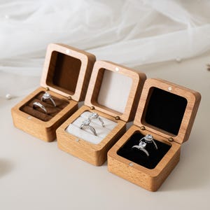 May include: Three square wooden ring boxes with hinged lids, each containing two silver-toned rings. The boxes feature interior velvet inserts in brown, white, and black, respectively, and are constructed from light-coloured natural wood.