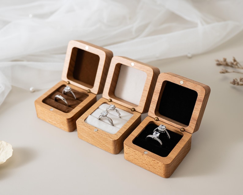 May include: Three square wooden ring boxes with hinged lids, each containing two silver-toned rings. The boxes feature interior velvet inserts in brown, white, and black, respectively, and are constructed from light-coloured natural wood.
