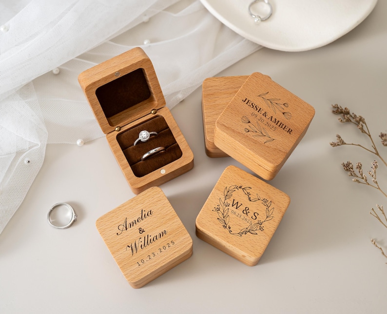 May include: Four square wooden ring boxes with hinged lids. One box is open, revealing a dark brown velvet interior holding two silver rings. The closed boxes feature laser-engraved names, dates, and floral designs including Jesse & Amber, Amelia & William, and W & S.