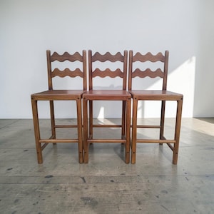 Pode incluir: Três cadeiras de madeira com acabamento castanho claro. Cada cadeira tem um assento quadrado, quatro pernas retas e um encosto com um design ondulado. As cadeiras estão lado a lado.