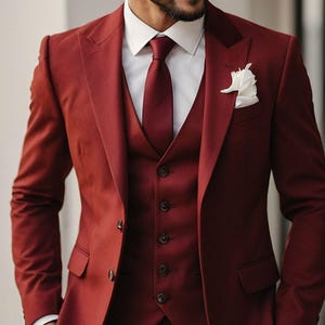 Può includere: Un abito bordeaux a tre pezzi con camicia bianca e cravatta bordeaux abbinata. L'abito comprende giacca, gilet e pantaloni. Un fazzoletto da taschino bianco aggiunge un dettaglio di contrasto. Un classico completo formale.