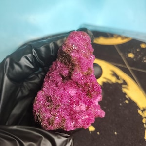 May include: A vibrant magenta crystalline mineral specimen with a rough, textured surface. The cluster features deep pink, sparkling crystal formations interspersed with patches of dark brown, earthy mineral deposits across its irregular shape.