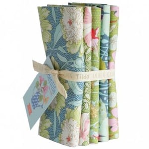 Könnte beinhalten: Ein Bündel gefalteter Stoffquadrate, gebunden mit einem cremefarbenen Band und einem Etikett. Die Stoffe zeigen florale Muster in Grün, Blau, Rosa und Weiß. Das Etikett trägt die Aufschrift "Gardenlife" und "Tilda Quilt Collection."