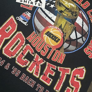 May include: Black t-shirt with a distressed graphic of the Houston Rockets NBA World Champions trophy. The design includes the team name, basketball, and an American flag-themed basketball. The text "'94 & '95 BACK TO BACK!" is also visible.