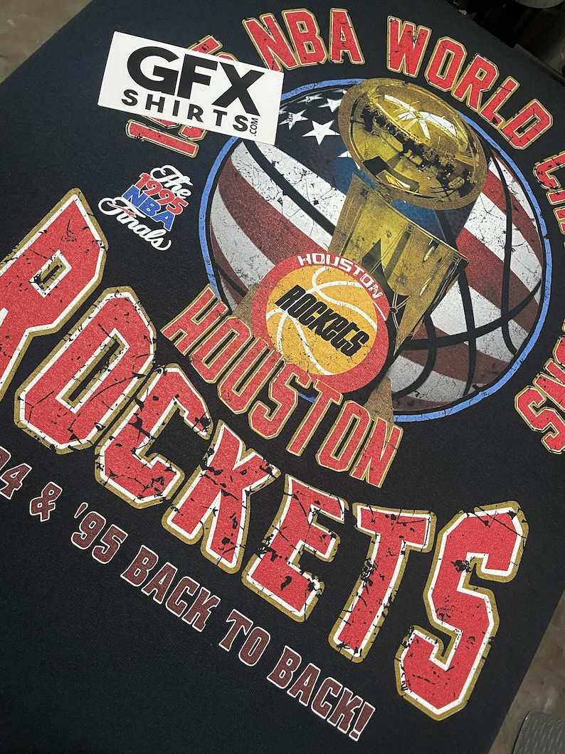 May include: Black t-shirt with a distressed graphic of the Houston Rockets NBA World Champions trophy. The design includes the team name, basketball, and an American flag-themed basketball. The text "'94 & '95 BACK TO BACK!" is also visible.