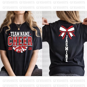 Op de afbeelding: Zwart T-shirt met "TEAM NAME CHEER 2026" in rode en witte letters en een pompom-afbeelding. De achterkant van het shirt heeft een strik met de naam "JASMINE" in het wit.