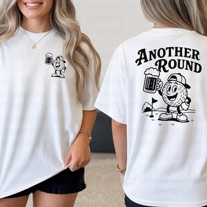May include: White t-shirt featuring a black and white cartoon golf ball character holding a beer mug. The front has a small graphic, while the back displays the text "ANOTHER ROUND" above the same character. A fun, casual tee.