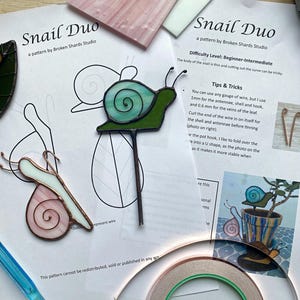May include: An overhead shot features stained glass snail art pieces and a pattern sheet titled "Snail Duo" by Broken Shards Studio. The image includes tools like pliers and a glass cutter, along with copper foil and glass pieces.