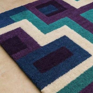 Puede incluir: Una alfombra con estampado geom&eacute;trico en tonos azules, turquesas, morados y crema. El dise&ntilde;o presenta cuadrados y zigzags. La alfombra est&aacute; hecha de un material suave y afelpado, adecuada para una sala de estar o dormitorio.