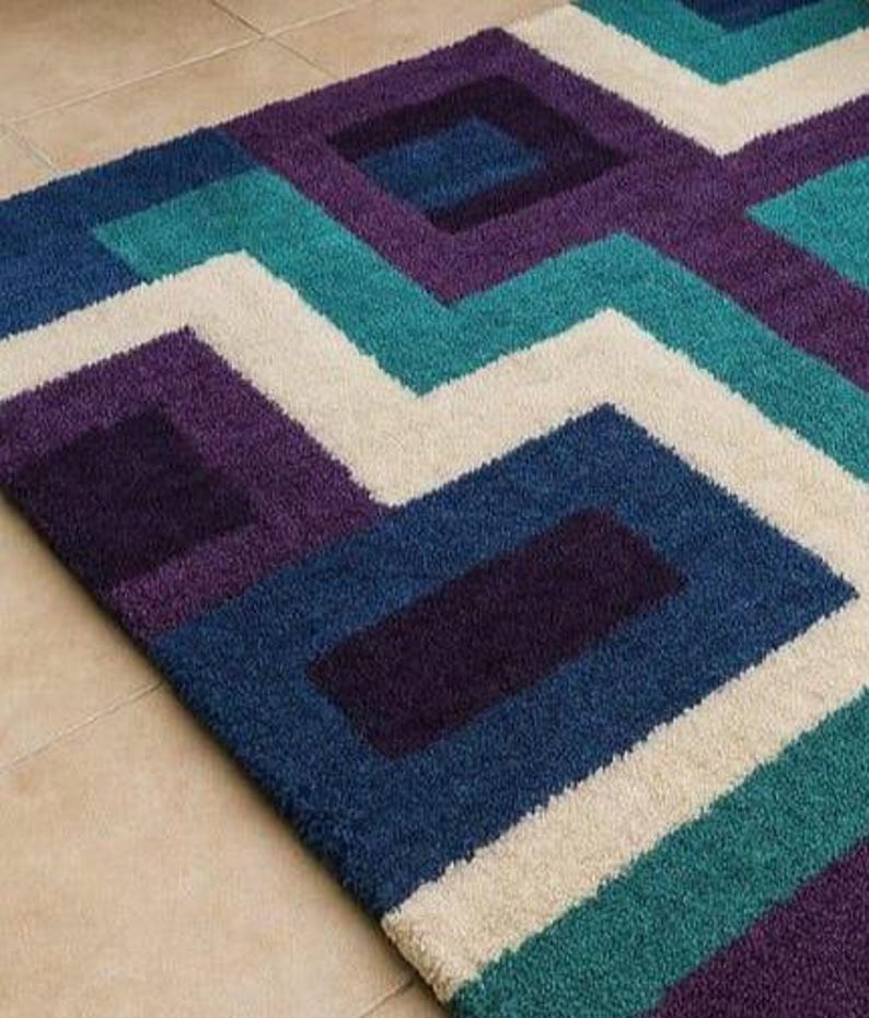 Puede incluir: Una alfombra con estampado geom&eacute;trico en tonos azules, turquesas, morados y crema. El dise&ntilde;o presenta cuadrados y zigzags. La alfombra est&aacute; hecha de un material suave y afelpado, adecuada para una sala de estar o dormitorio.