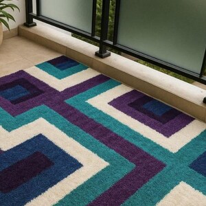 Puede incluir: Una alfombra rectangular con un patr&oacute;n geom&eacute;trico en tonos de azul, turquesa, morado y blanco. La alfombra presenta un dise&ntilde;o repetitivo de cuadrados y zigzags, creando una est&eacute;tica moderna. La alfombra est&aacute; hecha de un material suave y texturizado.