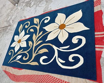 Puede incluir: Una alfombra rectangular con un fondo azul profundo y dos flores blancas grandes, cada una con un círculo central de color amarillo mostaza. El diseño incluye enredaderas y hojas de color beige que conectan los elementos florales.