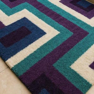 Puede incluir: Una alfombra con dise&ntilde;o geom&eacute;trico de l&iacute;neas y formas que se cruzan en tonos turquesa, morado, azul marino y crema. La alfombra est&aacute; hecha de un material suave y texturizado y est&aacute; colocada sobre un suelo embaldosado.