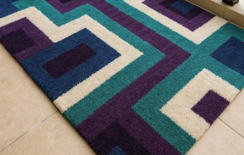 Puede incluir: Una alfombra con dise&ntilde;o geom&eacute;trico de l&iacute;neas y formas que se cruzan en tonos turquesa, morado, azul marino y crema. La alfombra est&aacute; hecha de un material suave y texturizado y est&aacute; colocada sobre un suelo embaldosado.