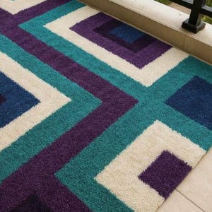 Puede incluir: Una alfombra rectangular con un patr&oacute;n geom&eacute;trico en verde azulado, morado, blanco roto y azul marino. El dise&ntilde;o presenta cuadrados y zigzags. La alfombra est&aacute; hecha de un material suave y afelpado, adecuado para uso en interiores.