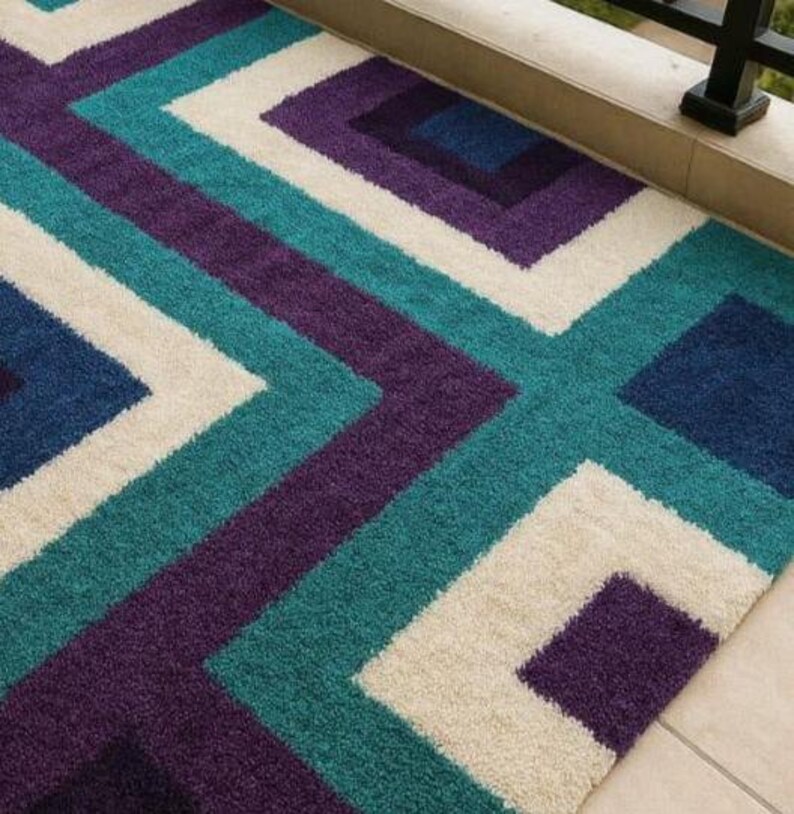Puede incluir: Una alfombra rectangular con un patr&oacute;n geom&eacute;trico en verde azulado, morado, blanco roto y azul marino. El dise&ntilde;o presenta cuadrados y zigzags. La alfombra est&aacute; hecha de un material suave y afelpado, adecuado para uso en interiores.