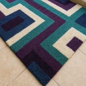 Puede incluir: Alfombra geom&eacute;trica con un patr&oacute;n de cuadrados y rect&aacute;ngulos en tonos turquesa, morado, azul marino y crema. El dise&ntilde;o de la alfombra crea un aspecto moderno. Est&aacute; hecha de un material suave y afelpado, ideal para una sala de estar o un dormitorio.