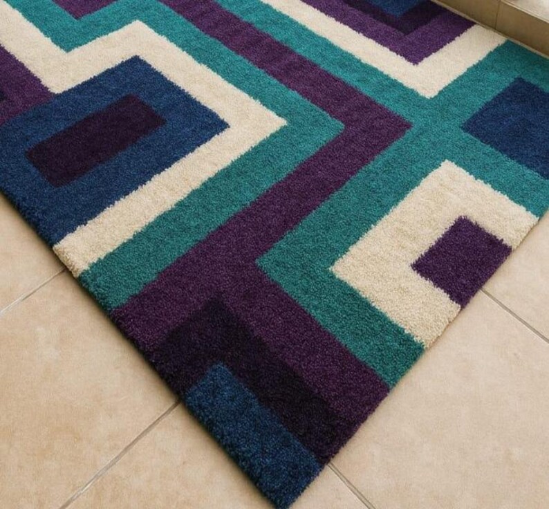 Puede incluir: Alfombra geom&eacute;trica con un patr&oacute;n de cuadrados y rect&aacute;ngulos en tonos turquesa, morado, azul marino y crema. El dise&ntilde;o de la alfombra crea un aspecto moderno. Est&aacute; hecha de un material suave y afelpado, ideal para una sala de estar o un dormitorio.