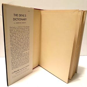 May include: An open book titled "The Devil's Dictionary" by Ambrose Bierce. The book's cover is black with white text, and the pages are off-white. The book is displayed on a white surface.