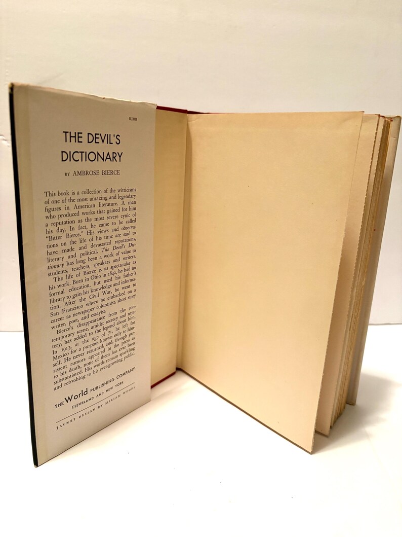 May include: An open book titled "The Devil's Dictionary" by Ambrose Bierce. The book's cover is black with white text, and the pages are off-white. The book is displayed on a white surface.