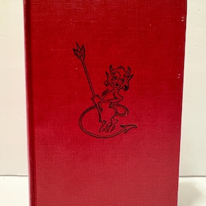 May include: A red hardcover book with a textured cover. The cover features a black illustration of a devil holding a pitchfork. The book is standing upright against a white background.