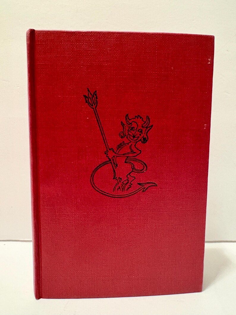 May include: A red hardcover book with a textured cover. The cover features a black illustration of a devil holding a pitchfork. The book is standing upright against a white background.
