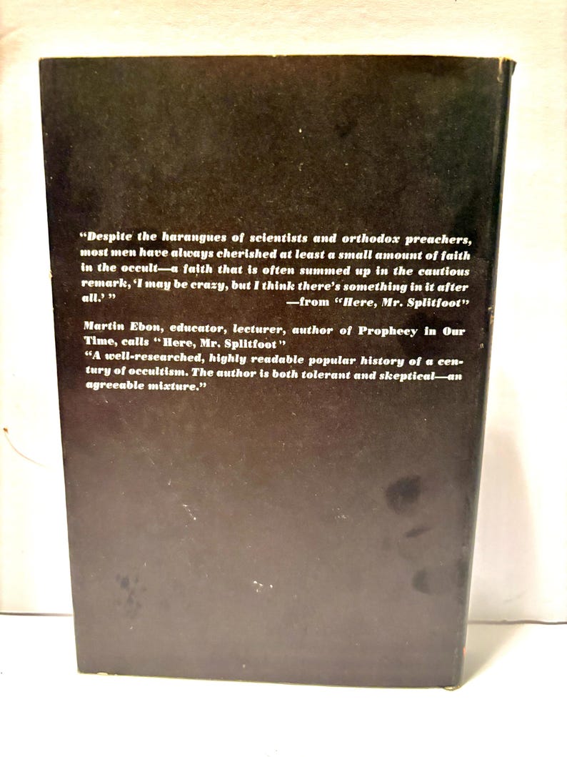 Puede incluir: La contraportada de un libro con un fondo marr&oacute;n oscuro. El texto blanco incluye citas e informaci&oacute;n del autor. El t&iacute;tulo del libro es "Here, Mr. Splitfoot". El libro trata sobre ocultismo.