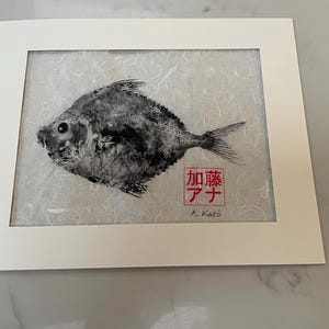 May include: A framed black and white fish print. The fish is centered on a textured, light-colored background. The print is in a white mat and frame. Japanese text and the name "A. Katō" are in the lower right corner.